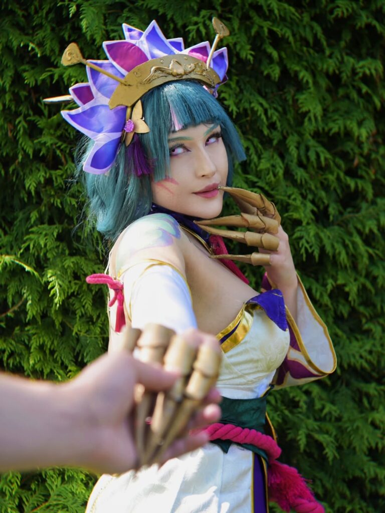 Cosplayer as Spirit Blossom Evelynn from League of Legends looking back over the shoulder while a hand in the foreground holds the golden claws, in front of a green hedge